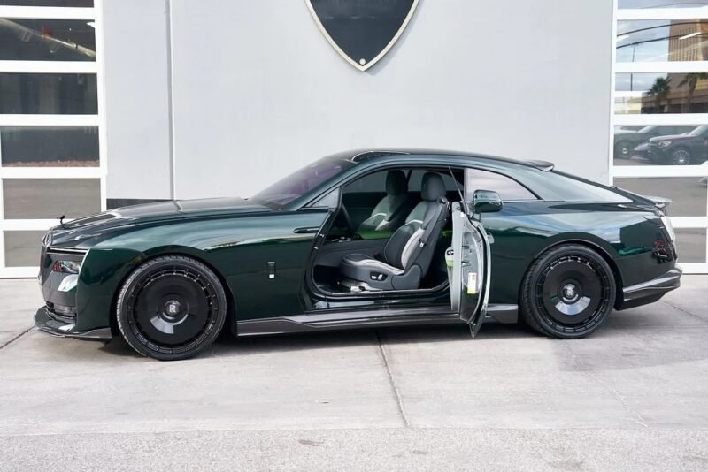 A dark green Rolls-Royce Spectre electric super coupe with its driver-side door open is parked in front of a modern building with large windows.