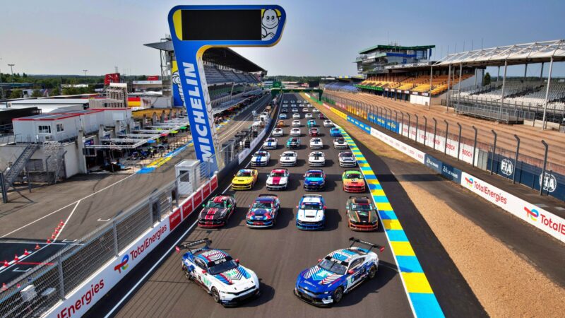 Race cars, including a Ford Le Mans contender, line up on the starting grid at a professional racing circuit in 2027, surrounded by grandstands, pit buildings, and banners echoing the iconic Ford vs Ferrari rivalry.