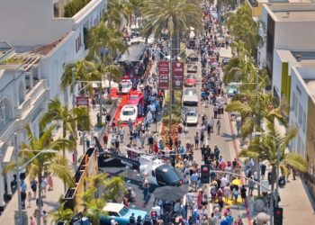 A large crowd gathers on a palm-lined street in Beverly Hills, with various cars and a helicopter on display during the June 15 Rodeo Drive Concours d’Elegance outdoor event.