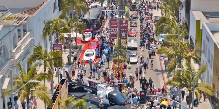 A large crowd gathers on a palm-lined street in Beverly Hills, with various cars and a helicopter on display during the June 15 Rodeo Drive Concours d’Elegance outdoor event.