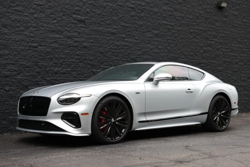 A silver luxury coupe from the best Bentleys, featuring sleek black wheels, is parked on a paved surface in front of a dark brick wall.