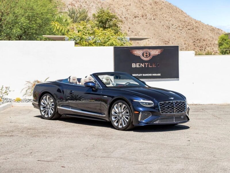A dark blue Bentley convertible, one of the best Bentleys, is parked outdoors near a white wall with a Bentley Rancho Mirage sign and a desert landscape in the background.