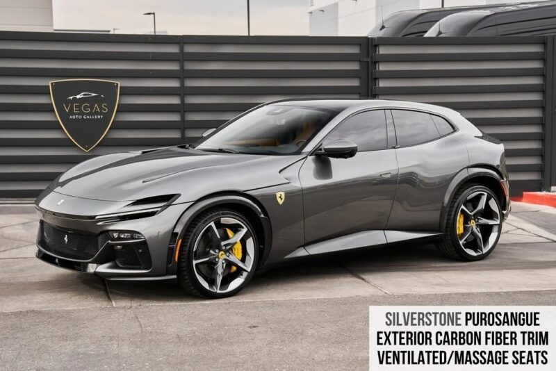 A grey Ferrari Purosangue SUV, one of the best Ferraris, is parked in front of a black fence with a Vegas Auto Gallery sign; features include carbon fiber trim and yellow brake calipers.
