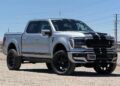 A silver Ford F-150 Shelby pickup truck with black racing stripes is parked on a dirt surface under a clear blue sky. Explore this striking example of Shelby Trucks—perfect if you're searching for a Ford F-150 for sale.