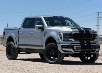 A silver Ford F-150 Shelby pickup truck with black racing stripes is parked on a dirt surface under a clear blue sky. Explore this striking example of Shelby Trucks—perfect if you're searching for a Ford F-150 for sale.
