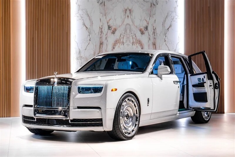 „White Rolls-Royce Phantom“ prabangus sedanas su chromo akcentais ir atviros galinės durys rodomos modernioje salone su vertikaliomis medžio plokštėmis ir marmurinėmis sienomis.