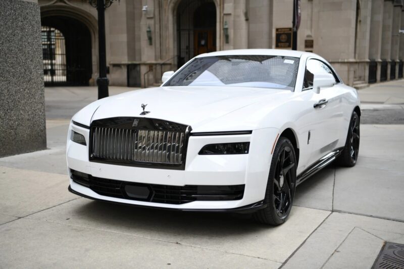 A white ultra-luxe Rolls-Royce Spectre electric super coupe is parked on a city street in front of a stone building with arched doorways.