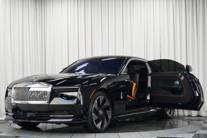 A black Rolls-Royce Spectre Ultra-Luxe sedan with a distinctive grille and orange-accented interior is parked indoors, its rear passenger door open.