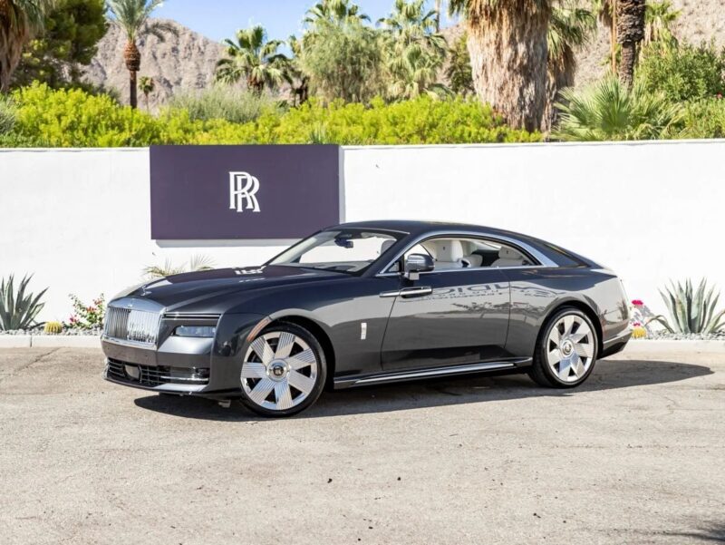 An ultra-luxe, dark gray Rolls-Royce Spectre electric super coupe is parked outdoors in front of a white wall with a Rolls-Royce logo, surrounded by palm trees and lush greenery.