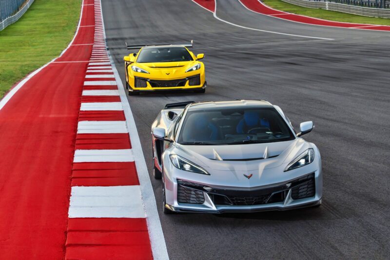 A silver Chevrolet Corvette ZR1X supercar, boasting 1,250-HP, leads a yellow Corvette on a race track with red and white curbs.