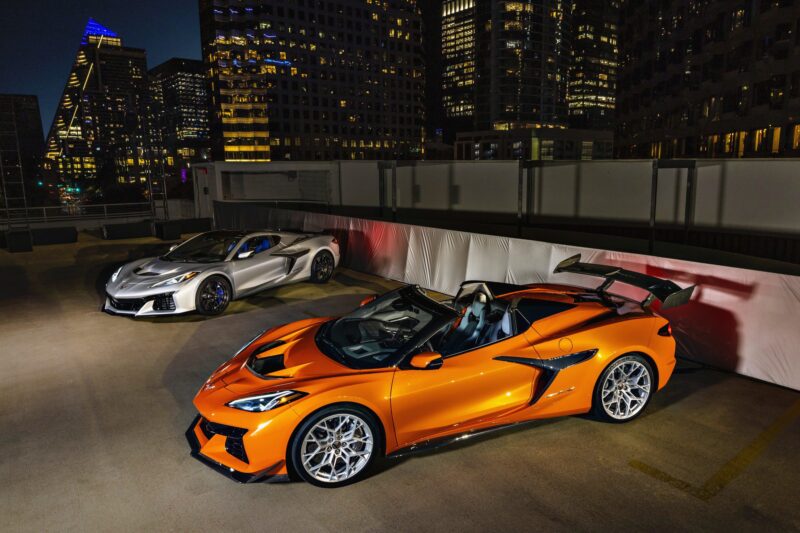 Two supercars, one orange and one silver, are parked on a rooftop at night, city buildings illuminated in the background.