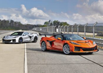Two supercars—a silver and an orange Chevrolet Corvette ZR1X with 1,250-HP—are parked on a racetrack next to a fenced barrier under a partly cloudy sky.