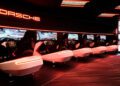 A row of Porsche-branded racing simulator seats with steering wheels and screens lines this modern, neon-lit gaming center at One Porsche Drive in Atlanta.