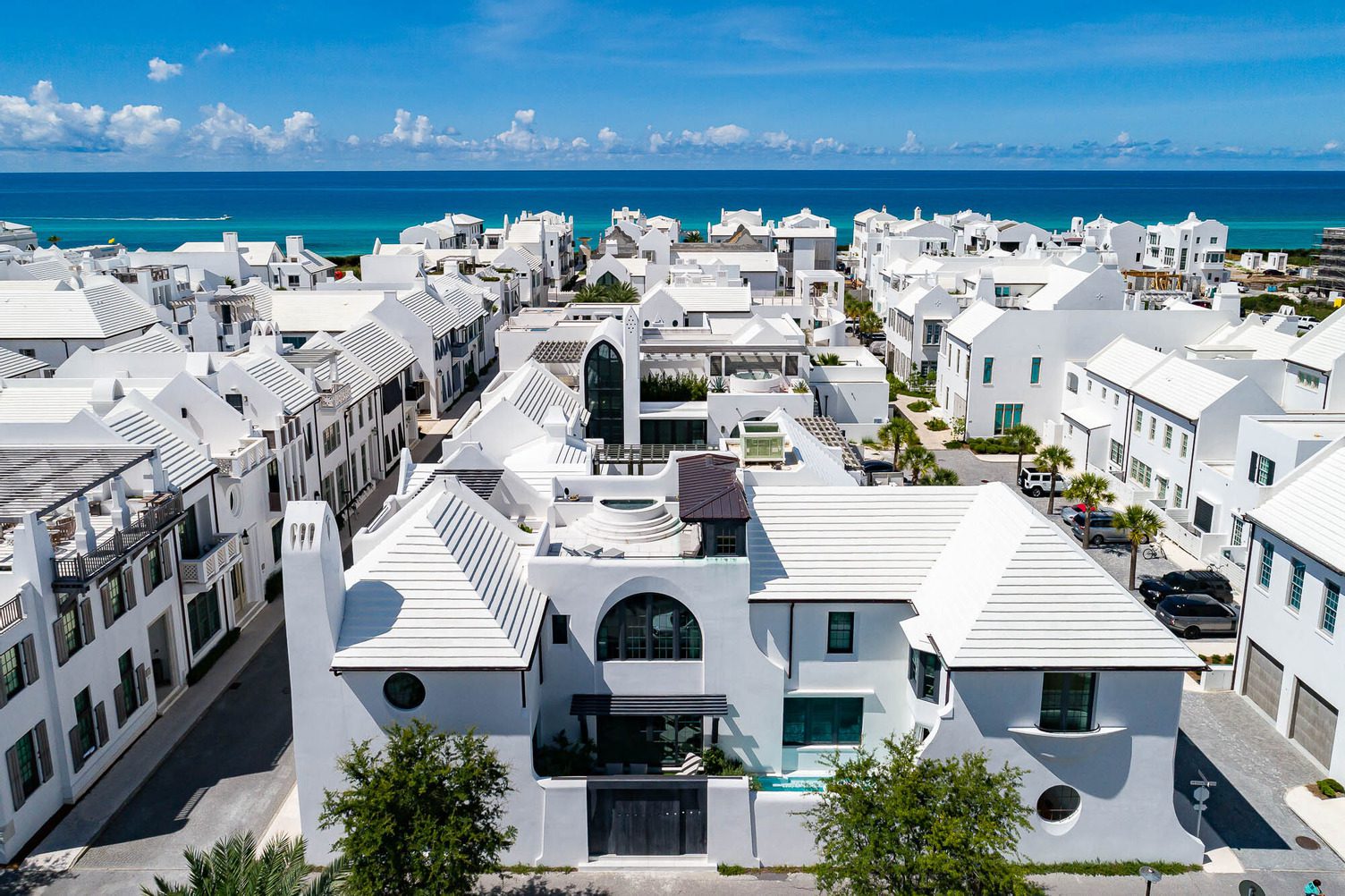 Aerial view of Alys Beach’s coastal neighborhood with white modern houses and rooftops, blue ocean in the background, and a few Cars of 30A gleaming under the sunny sky.
