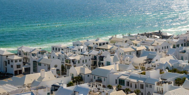 White-roofed, modern buildings of Alys Beach are clustered along a coastline with turquoise ocean waves, sunlight reflecting on the water, and hints of the Art of the Drive along scenic 30A.