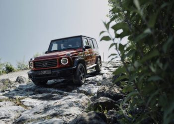 A red Mercedes-Benz G-Class SUV is parked on rocky, uneven terrain with greenery and clear sky in the background, capturing the spirit of adventure that drives demand for the G-Class Experience Program.