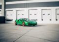 A green Ferrari 296 GTS with gold wheels, a modern tribute to classic design, is parked on a concrete surface in front of white industrial garage doors.