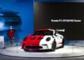 A Porsche 911 GT3 RS with red and white livery is displayed on a circular platform at the Porsche Experience Center Toronto; a person stands nearby and a screen reads "Porsche 911 GT3 RS PEC Toronto.