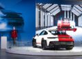 A white and red 911 GT3 RS is displayed on a circular platform indoors at the Porsche Experience Center, with a mannequin in racing gear and a large screen showcasing a similar car in the background.