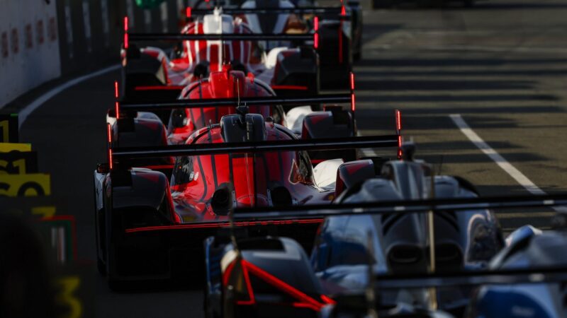 Multiple Le Mans prototype race cars based on the Hypercar platform are lined up closely together in a pit lane, viewed from the rear, with rear wing lights illuminated.