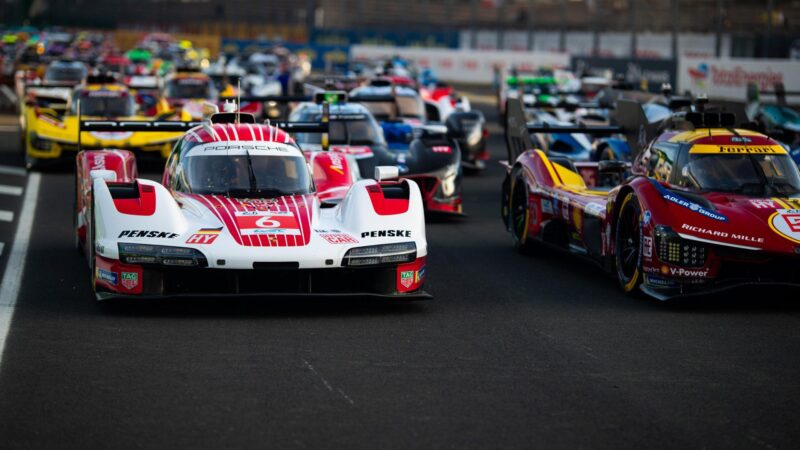 Multiple race cars line up at the starting grid on a racetrack, with a white Porsche Hypercar and a red Ferrari leading the pack in this FIA WEC event.