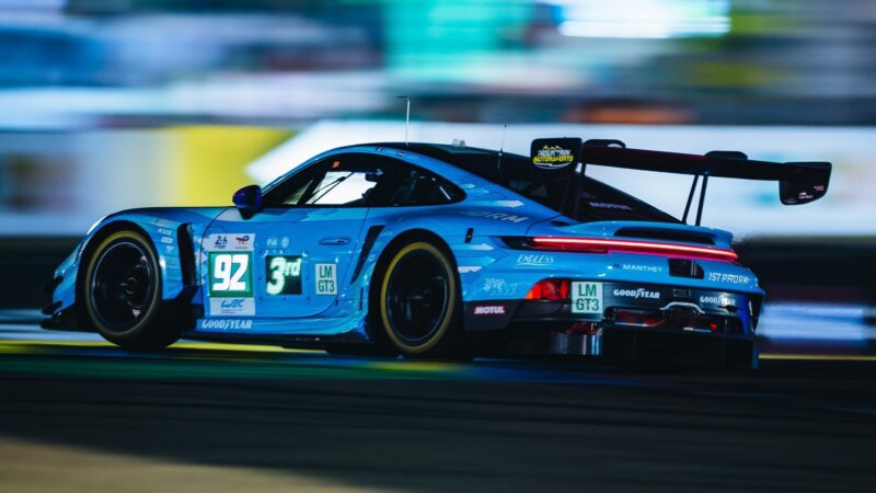 A blue Porsche race car with number 92 speeds on a racetrack at night, displaying sponsor logos and a prominent rear wing, as Porsche denies podium sweep while Ferrari clinches historic third straight Le Mans victory.