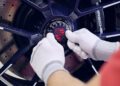 Close-up of gloved hands installing a Porsche Italia center lock cap onto a black alloy wheel, with the brake disc visible behind.