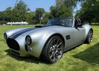A silver convertible sports car with black stripes is parked on grass, with trees and tents in the background under a clear sky.