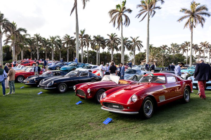 A row of classic cars, including an iconic Enzo Ferrari, is displayed on a lawn with people walking around and palm trees in the background.