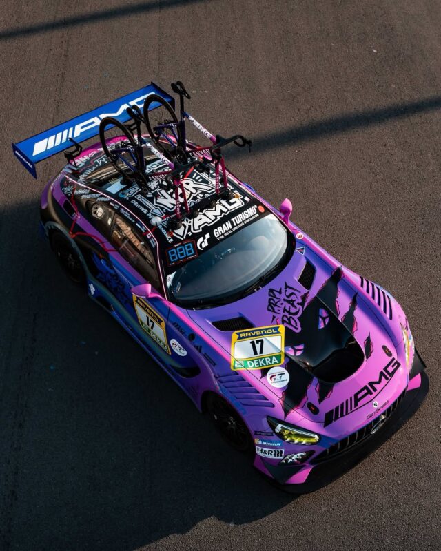 A purple and black Mercedes-AMG racing car with sponsor logos and two Canyon Aeroad Road Bikes mounted on the roof is seen parked on a sunlit road.