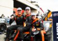 Three race car drivers in black and orange suits sit and pose together in front of an LMP2 race car at a racing event, smiling and wearing sponsor hats.