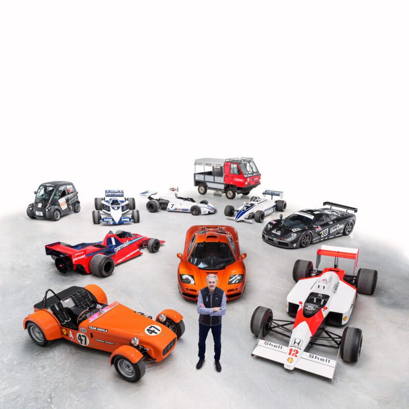 A person stands with arms crossed in front of a group of diverse racing and specialty cars, including models from Gordon Murray Special Vehicles, arranged in a semi-circle on a plain surface at Monterey Car Week.