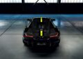 A black Lamborghini sports car with yellow racing stripes is parked indoors on a concrete floor, viewed from behind under soft industrial lighting.
