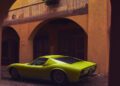 A bright green Lamborghini in Verde Scandal is parked on a cobblestone street beneath arches of a yellow building with wooden shutters and a mural.