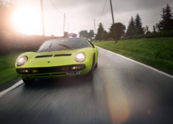 A green Lamborghini in stunning Verde Scandal cruises along a wet country road lined with trees and power lines, set against a cloudy sky—a story of speed and style.
