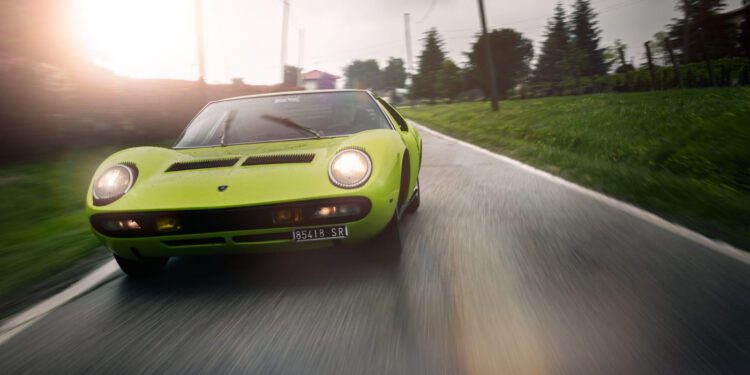 A green Lamborghini in stunning Verde Scandal cruises along a wet country road lined with trees and power lines, set against a cloudy sky—a story of speed and style.