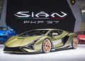 A Lamborghini Sián FKP 37 in metallic green is displayed on a stage at Monterey Car Week, with the model name and logo behind it. Blue and red Lamborghini Few-Off cars are visible in the background.