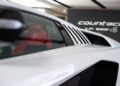 Close-up side view of a white Lamborghini Countach with visible red seat and Lamborghini logo, captured during Monterey Car Week, with a "Countach" display sign in the background.