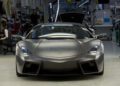 A matte gray Lamborghini Few-Off Legacy sports car is parked inside an automotive factory, with various equipment and workers visible in the background.