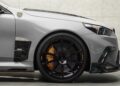 Close-up of the front side of a gray sports car, showing the wheel, brake caliper, and part of the headlight against a light-colored wall background.