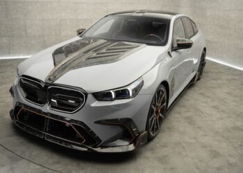 A modern luxury sedan with a custom carbon fiber hood and trim, displayed in a minimalistic showroom with concrete walls and floor.