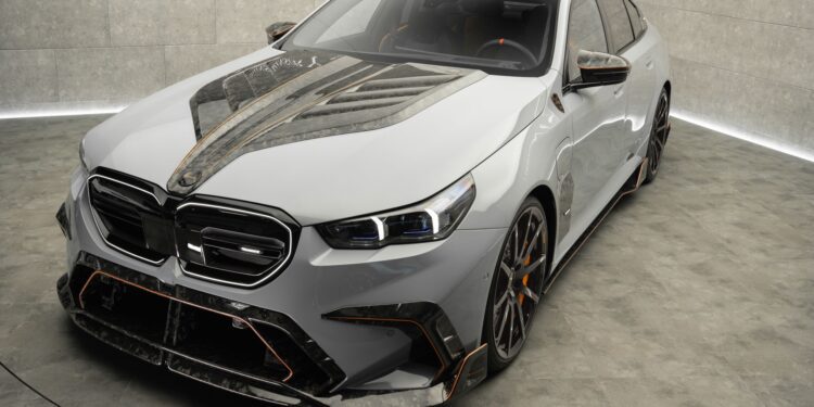 A modern luxury sedan with a custom carbon fiber hood and trim, displayed in a minimalistic showroom with concrete walls and floor.