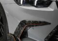 Close-up of the front left section of a silver sports car, showing the headlight, part of the front wheel, and a carbon fiber and orange-accented aerodynamic splitter.