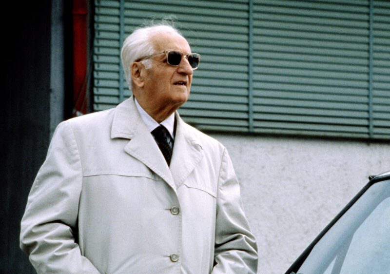 An older man wearing sunglasses and a light-colored trench coat stands outdoors near a building with green shutters and a car, evoking Enzo Mattioli Ferrari’s timeless style and hinting at Enzo’s Lasting Legacy.