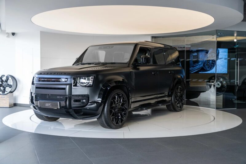 SUV Matte Black Land Rover Defender SUV, one of the most prominent unique cars, is shown on a circular platform in a modern showroom with reflective surfaces and bright lighting.