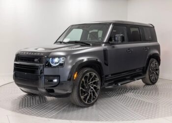 A matte black Land Rover Defender SUV with custom wheels, black trim, and Overfinch branding is parked in a white, well-lit showroom—a true unique garage creation for fans of custom cars.