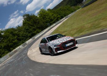 A camouflaged car with German license plates drives through a curve on a racetrack surrounded by grass and trees under a partly cloudy sky.