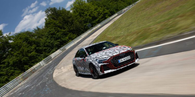 A camouflaged car with German license plates drives through a curve on a racetrack surrounded by grass and trees under a partly cloudy sky.