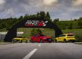 Three brightly colored cars—yellow, red, and yellow—are parked in front of the Salzburg Ring racetrack entrance on a sunny day with green trees in the background.