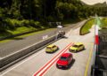 Three compact cars—two yellow and one red—are parked side by side on a race track next to a pit lane, with green hills and trees in the background.
